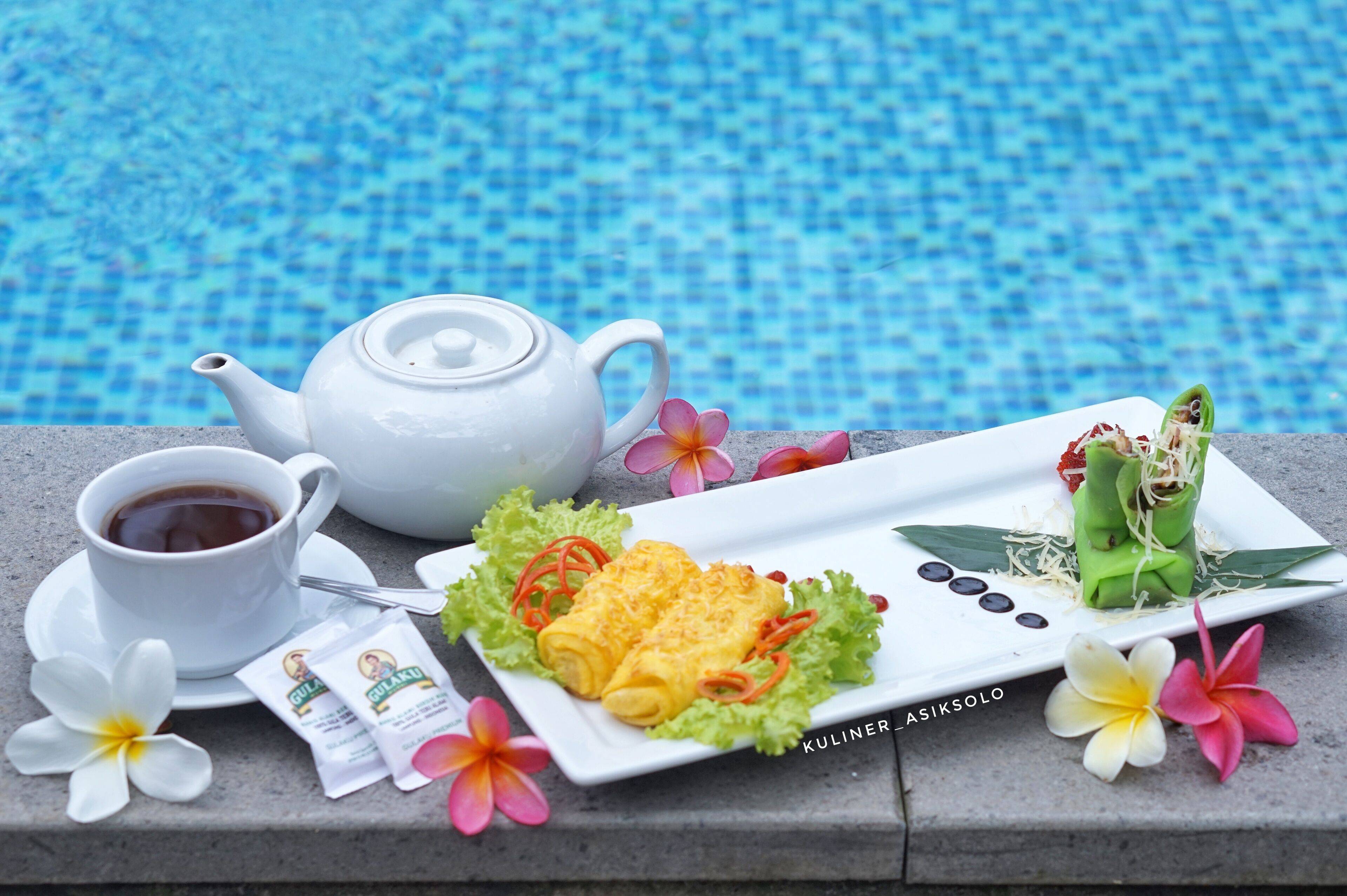 A table with a white teapot, a cup of tea, a plate of food, and some flowers by a pool.