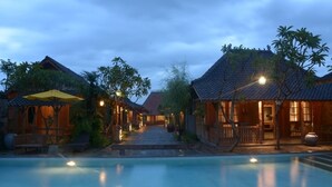 A poolside area with traditional architecture, a clear pathway leading to a building, and a calm water surface reflecting the lights.