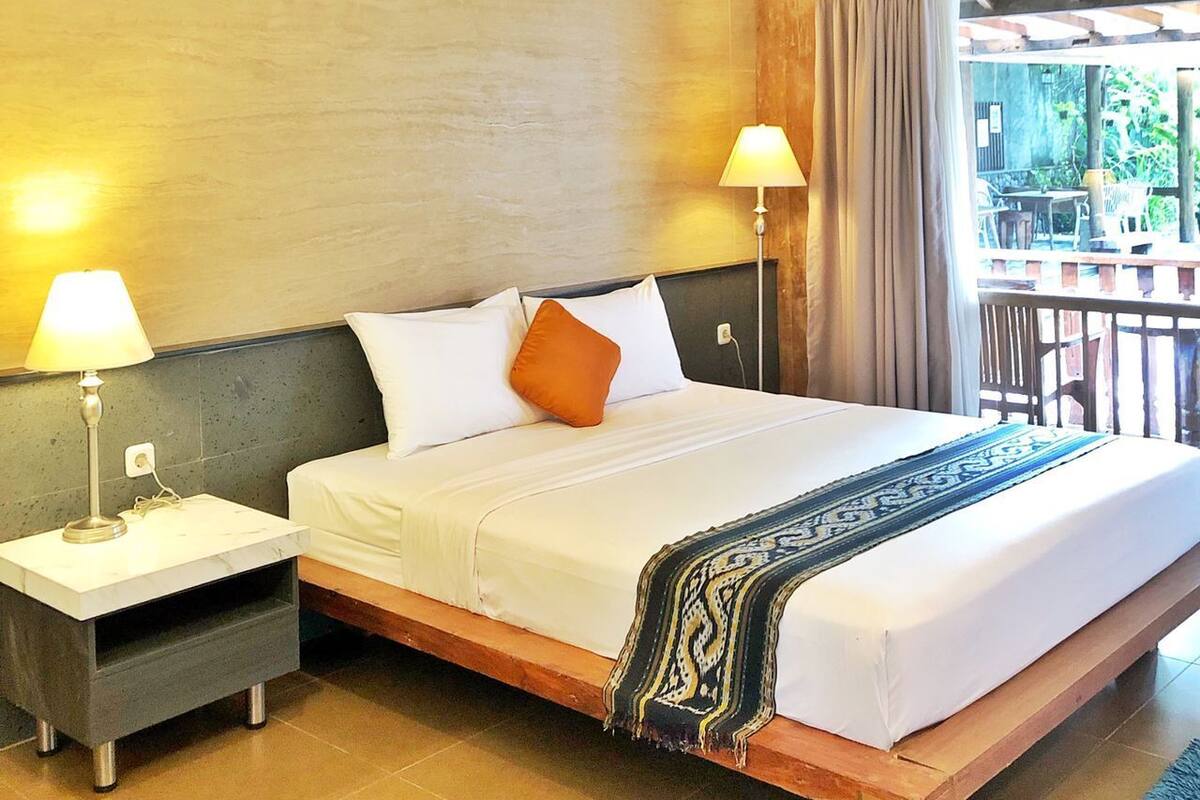 A hotel room with a large bed, two bedside lamps, a wooden headboard, and a view of the outdoors through a balcony.