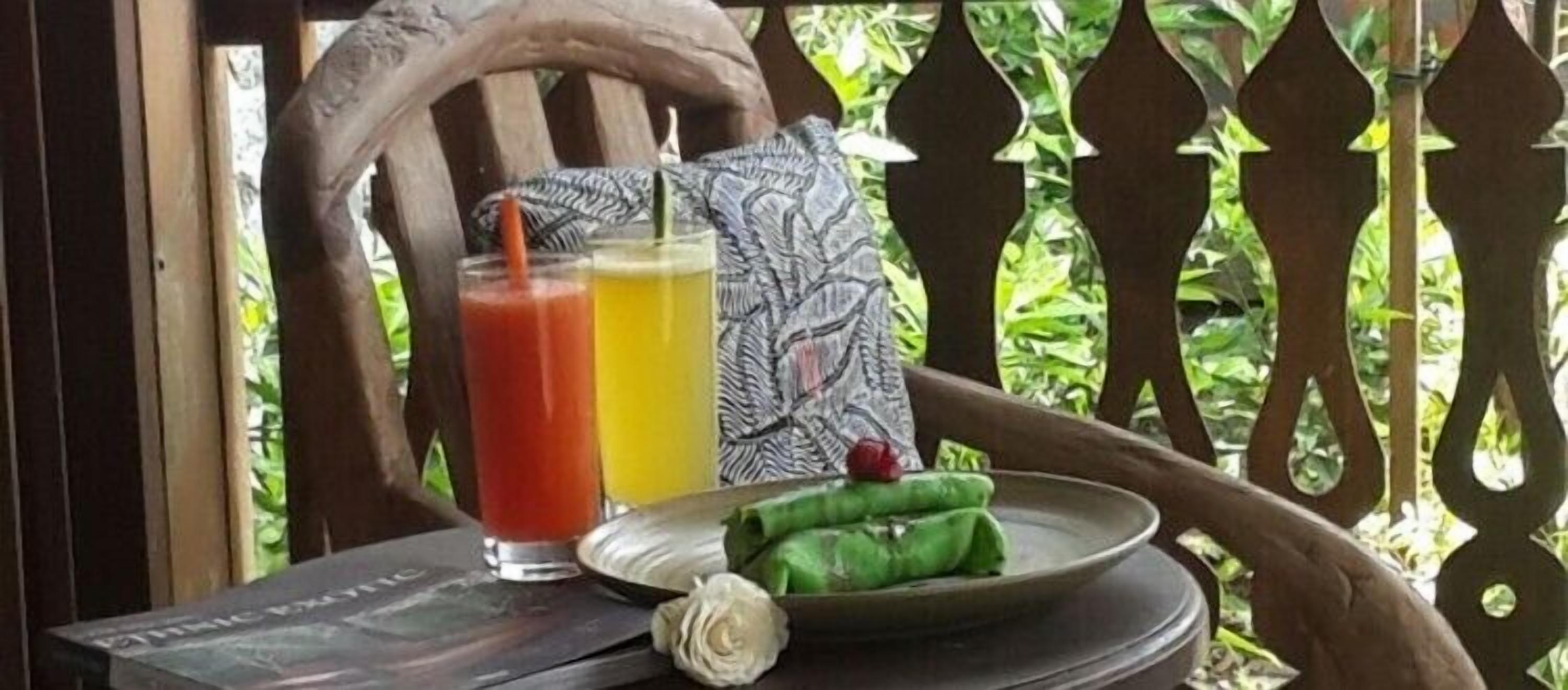 A table with two glasses of juice and a plate with a wrapped item, set against a wooden chair and a decorative fence.