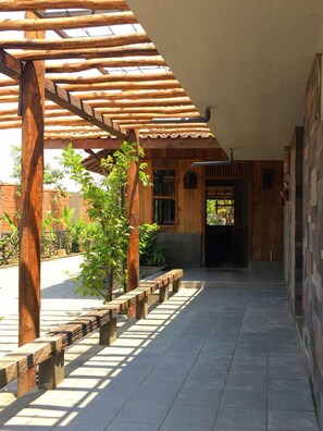 Un espacio exterior techado con vigas de madera y piso de baldosas, que conduce a la entrada de un edificio.