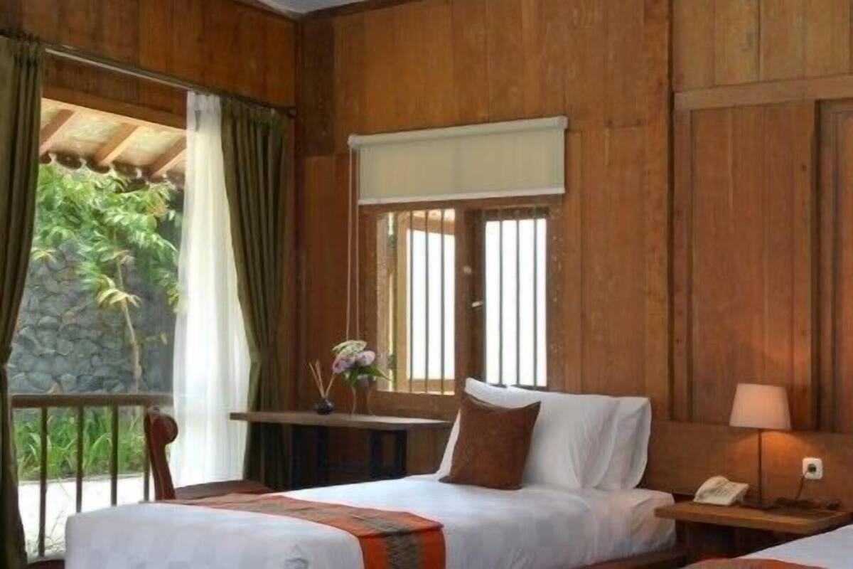 A room with two beds, wooden walls, a window with blinds, a nightstand with a lamp, and a view of greenery outside.