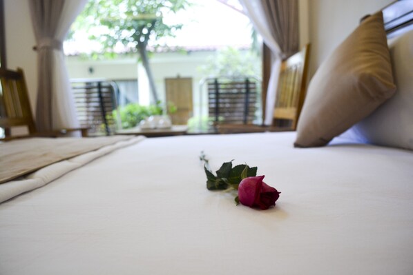 A bed with a single rose, a view of a balcony with plants, and a window with curtains. - Ngoc Viet Bungalow (Phu Quoc)