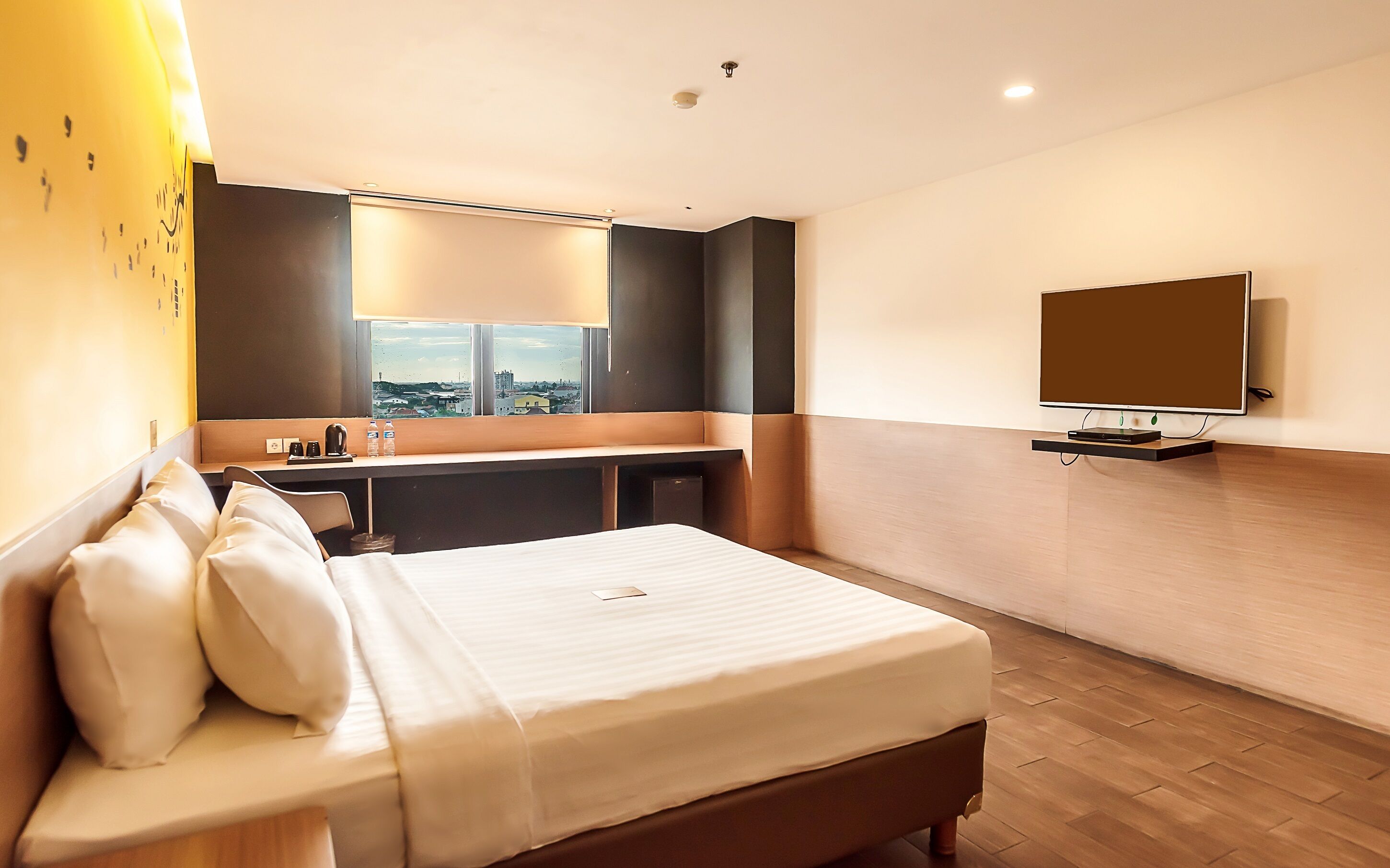 A hotel room with a bed, a desk with a chair, a TV mounted on the wall, and a window with a city view.