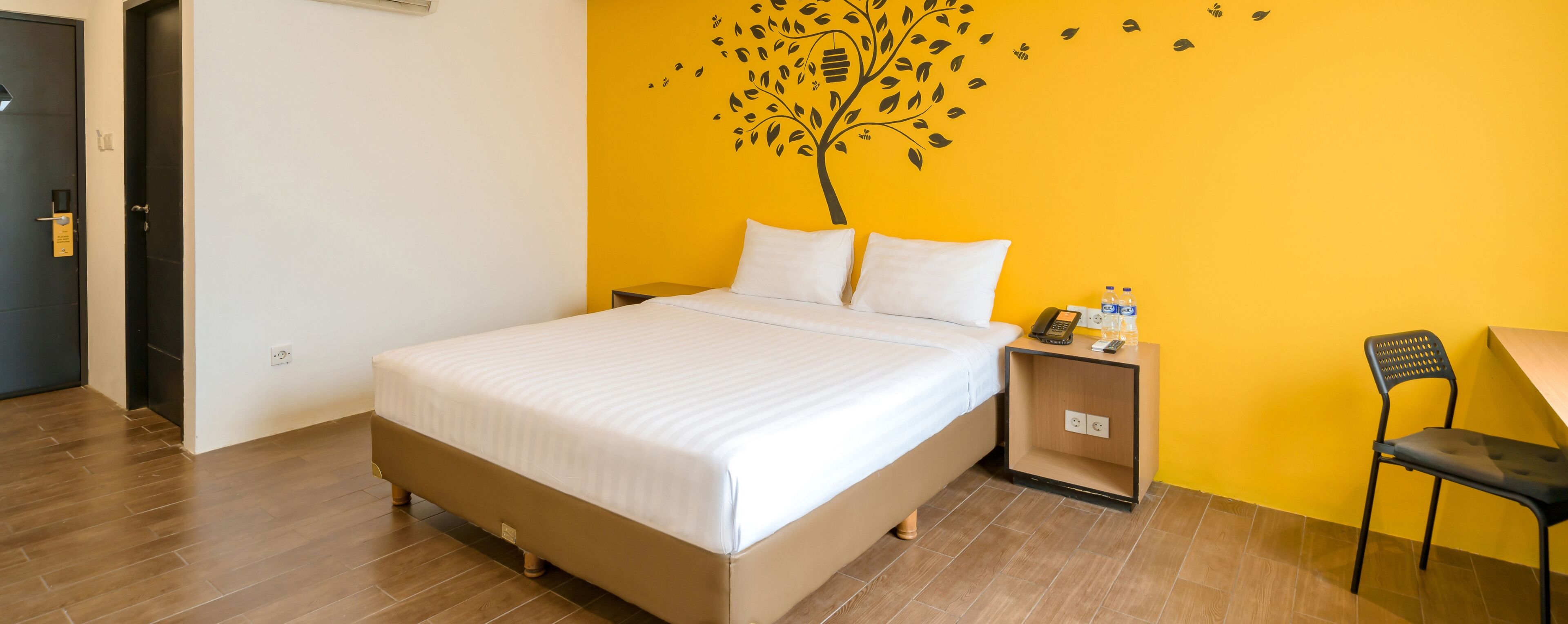 Hotellihuone, jossa on sänky, yöpöytä, jossa on puhelin ja lasi vettä, tuoli ja seinä, jossa on puukuva.