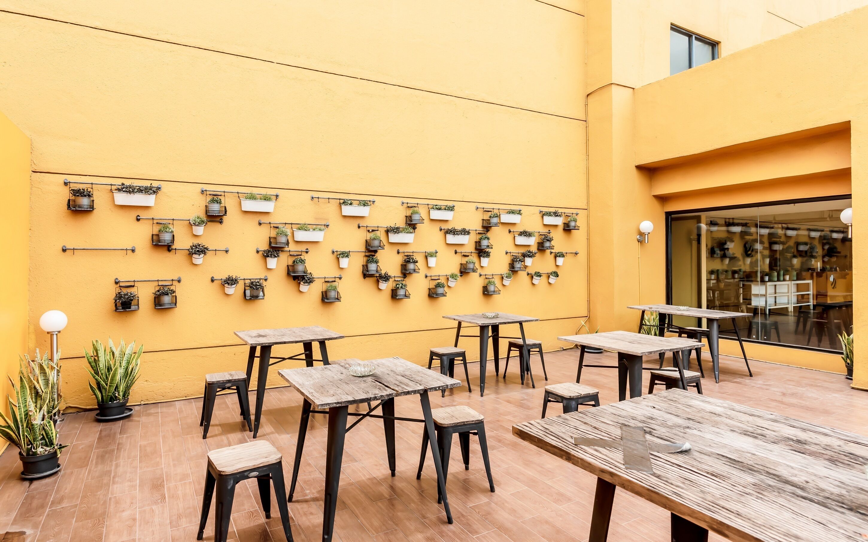 An outdoor seating area with multiple tables and chairs, a yellow wall with potted plants, and a glass door leading inside.