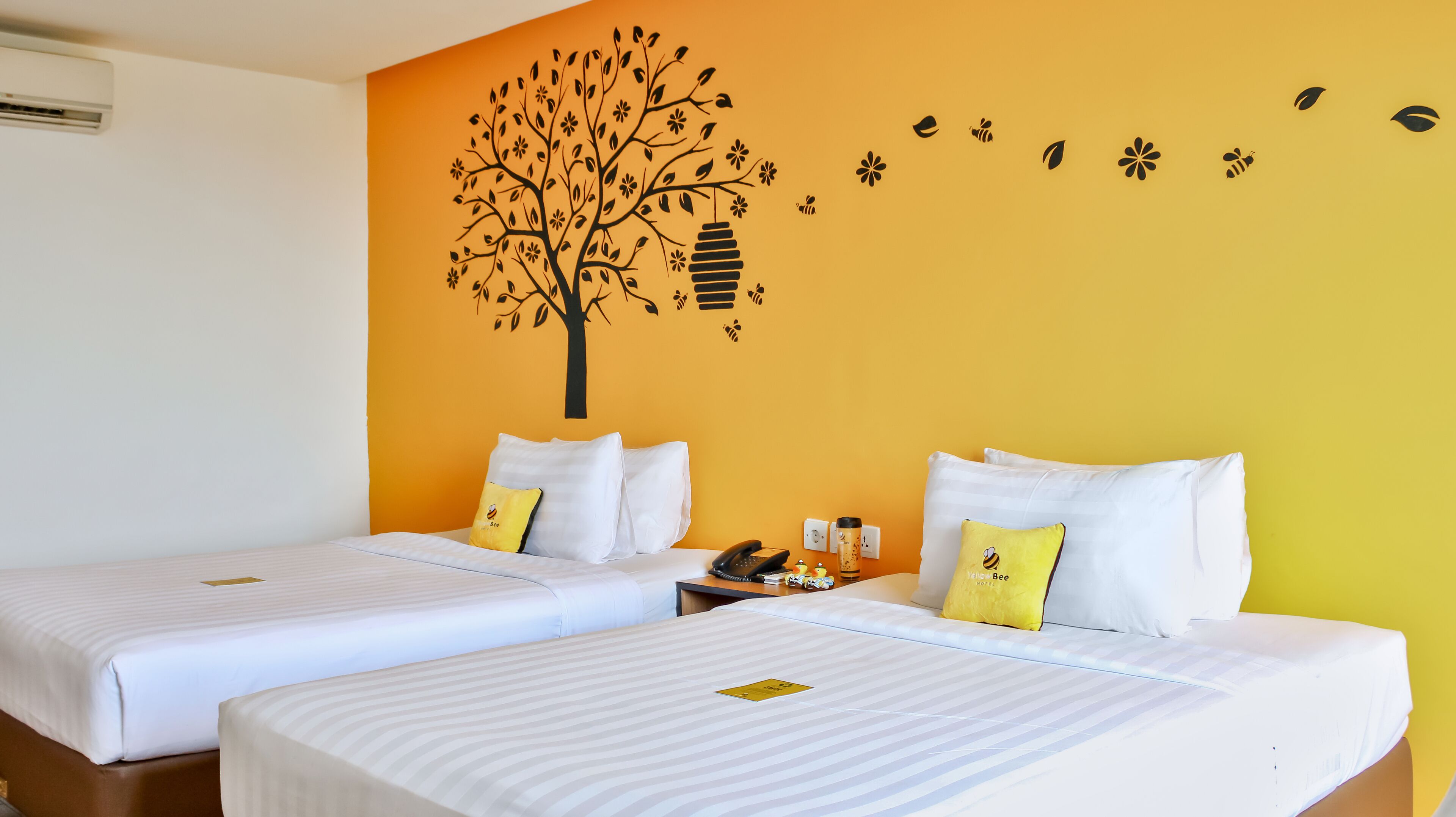 Habitación de hotel con dos camas, ropa de cama blanca, un mural de un árbol en la pared y una pared amarilla.