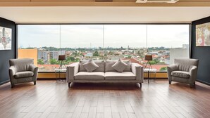 Habitación amplia con un ventanal grande que ofrece vistas a la ciudad, un sofá, dos sillones y una mesita con lámparas.