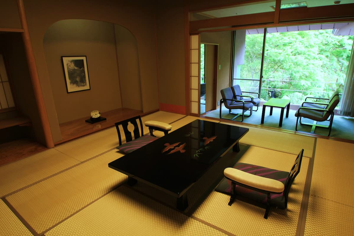 Una habitación con esteras tatami, una mesa baja y sillas, y un fusuma que da a un balcón con vistas a árboles.
