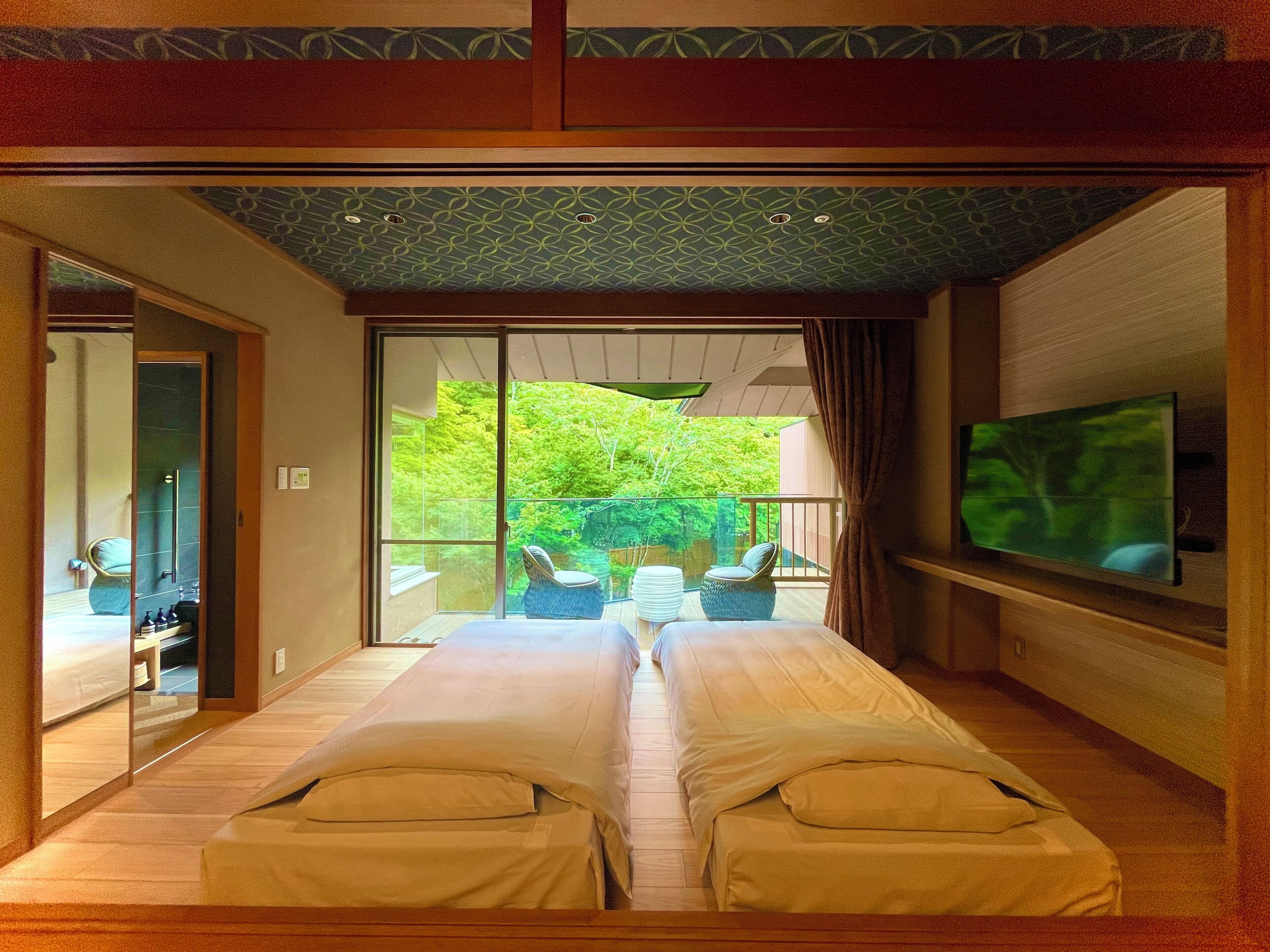 A bedroom with a large bed, a TV, and a sliding glass door leading to a balcony with outdoor furniture.