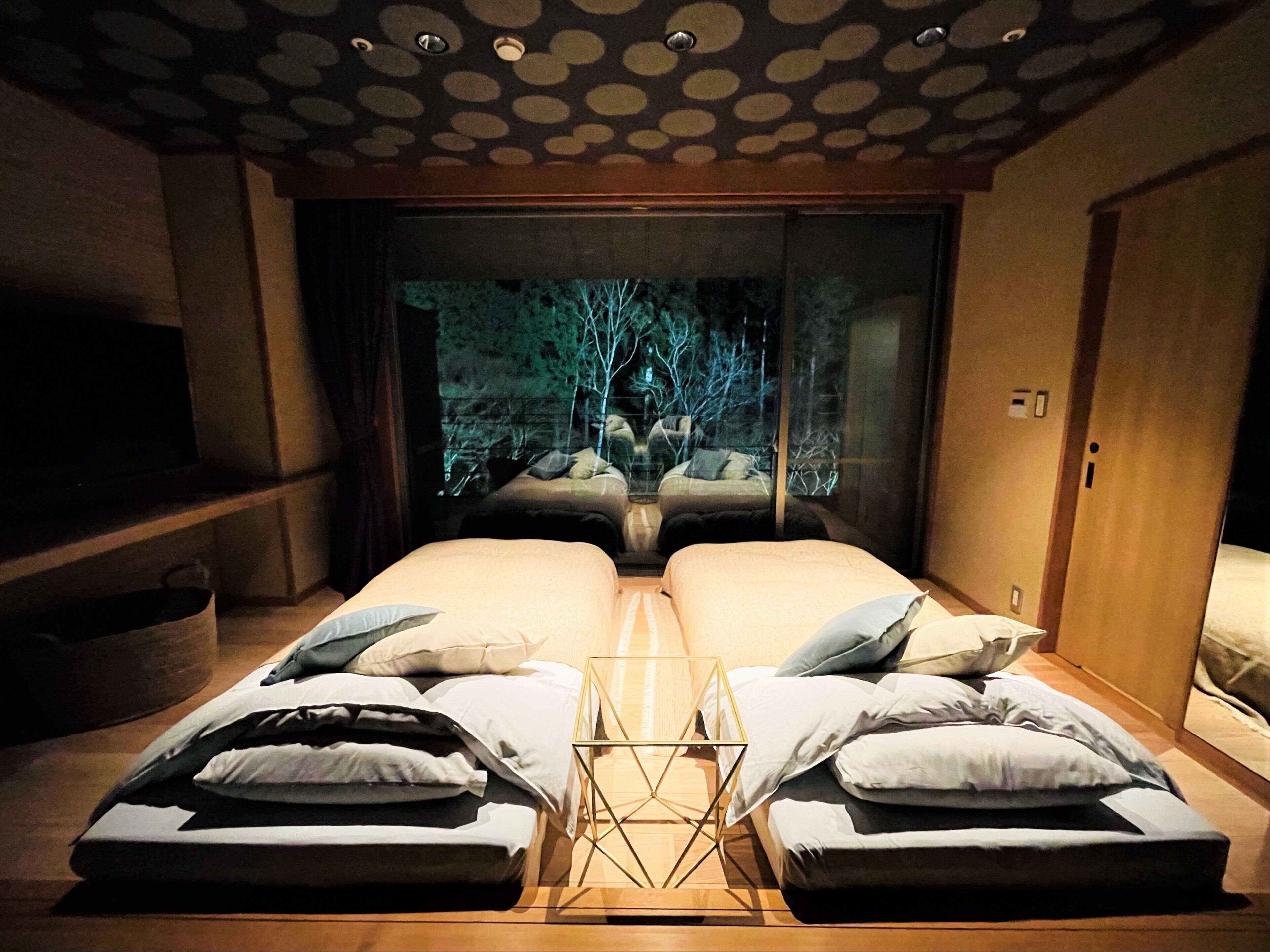 A room with two beds, a small table, and a view of trees through a large window.
