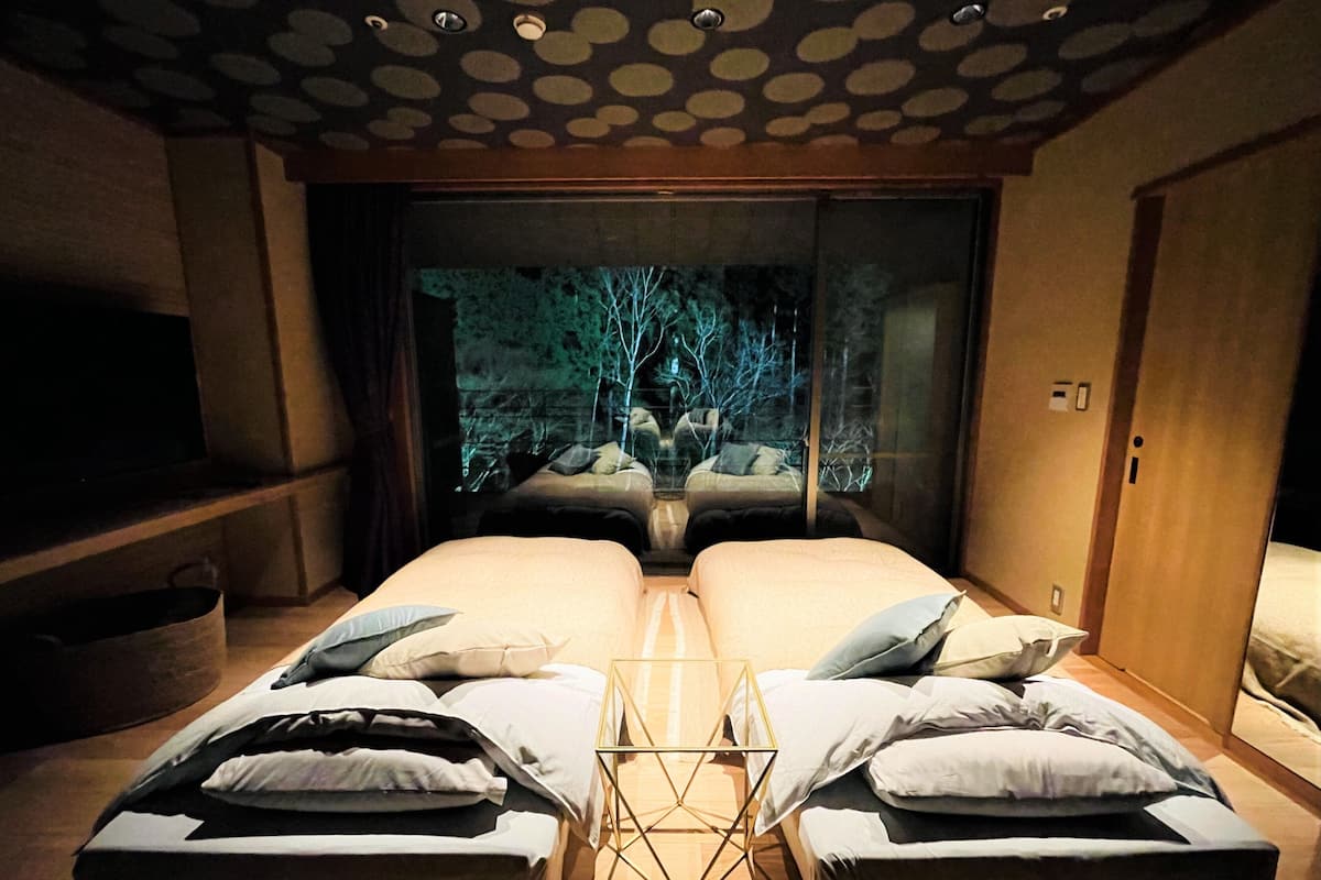 Una habitación con dos camas, una mesita y vistas a árboles a través de un amplio ventanal.