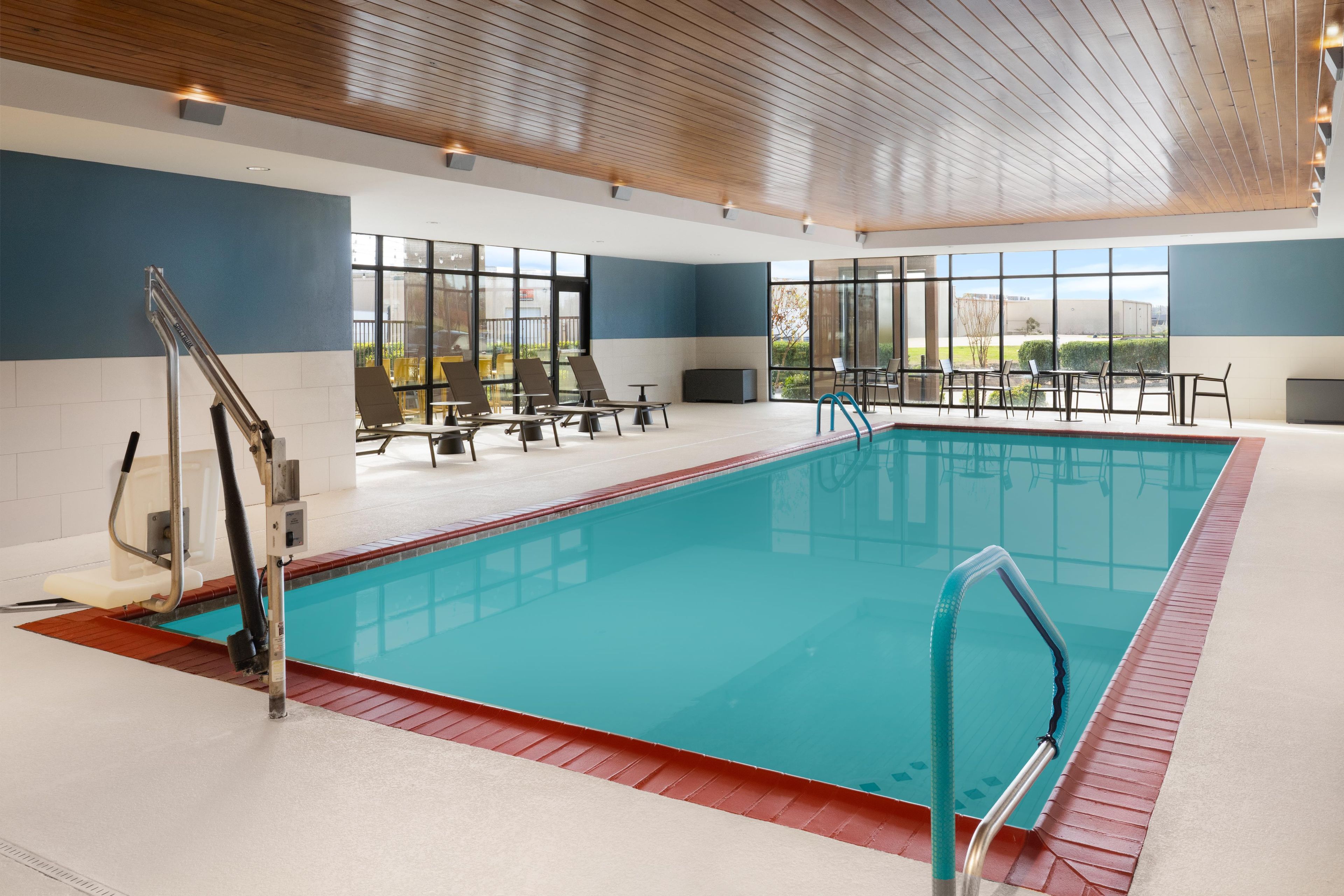 An indoor swimming pool with clear glass walls, a wooden ceiling, and a poolside seating area.