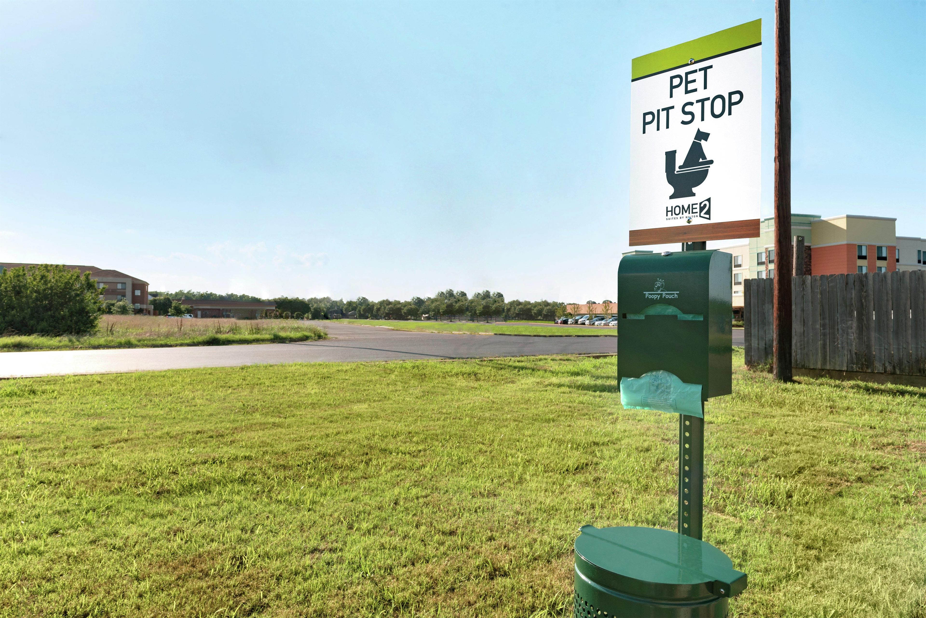 A pet waste station with a sign indicating a pet pit stop, a green trash bin, and a building in the background.