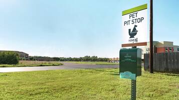 A pet waste station with a sign indicating a pet pit stop, a green trash bin, and a building in the background.
