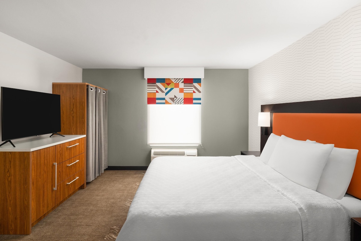 A hotel room with a bed, a TV, a wardrobe, and a window with a geometric curtain.