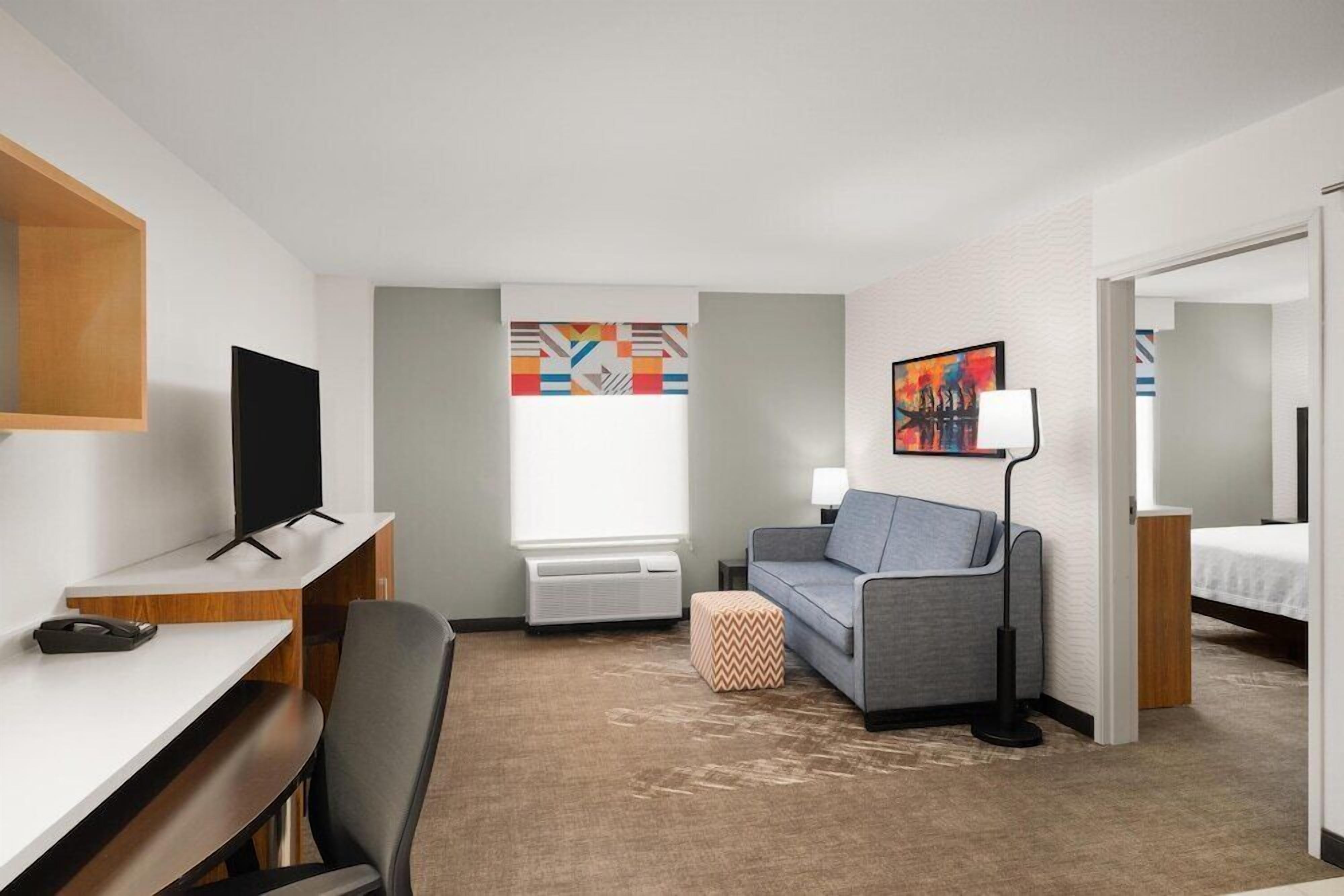 A hotel room with a desk, a grey sofa, a TV, a lamp, a bed, and a window.