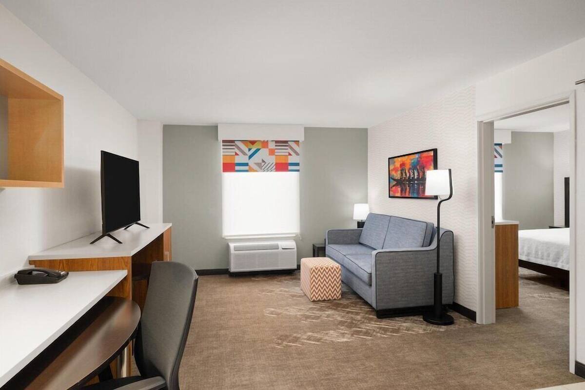 Habitación de hotel con escritorio, sofá gris, televisor, lámpara, cama y ventana.