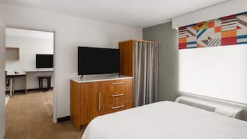 A hotel room with a bed, a TV on a wooden cabinet, a desk with a chair, and a colorful abstract wall art.