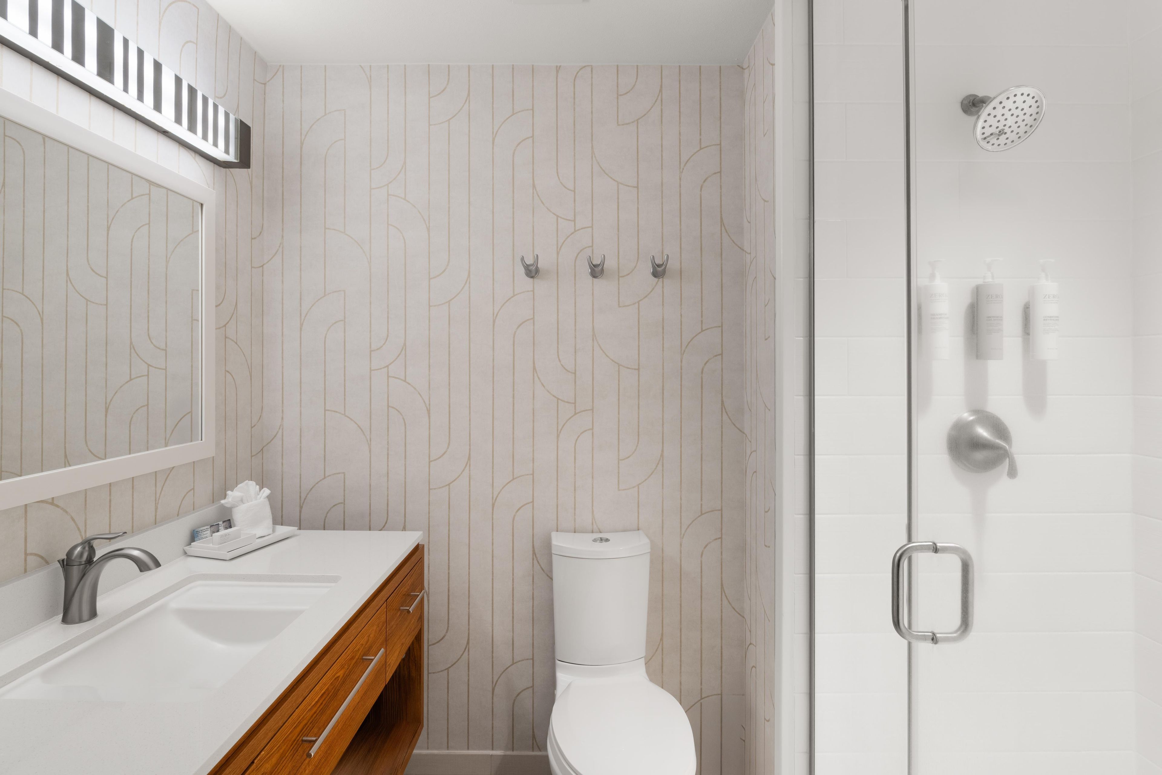 A modern bathroom with a white sink, mirror, and toilet. There is a shower area with a handheld showerhead and a towel rack.