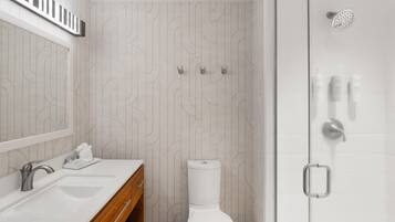 A modern bathroom with a white sink, mirror, and toilet. There is a shower area with a handheld showerhead and a towel rack.