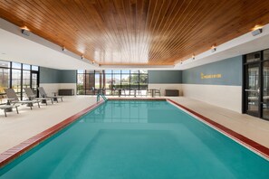 Una piscina con sillones de descanso y mesas, techo de madera y ventanas amplias.