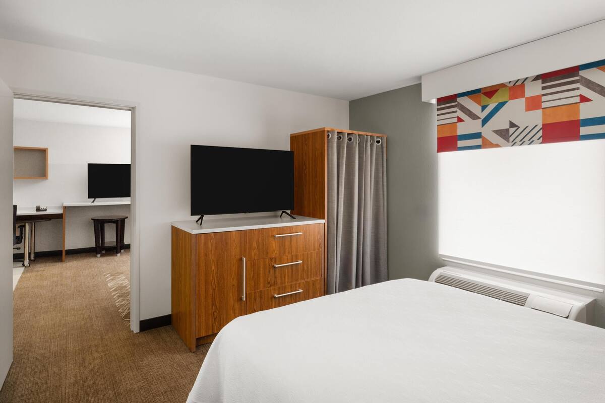 Habitación de hotel con cama, armario de madera, escritorio con silla, televisor y ventana con cortinas.