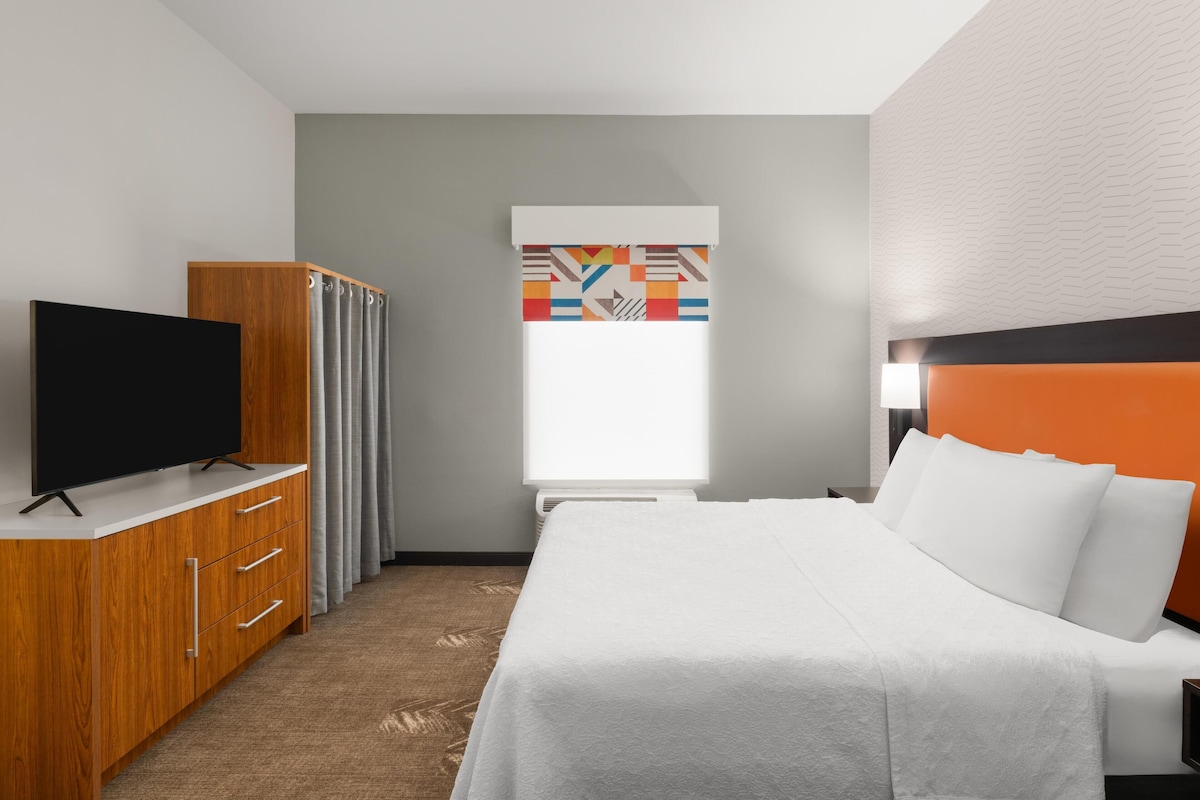 A hotel room with a bed, a TV on a wooden cabinet, a window with curtains, and a patterned wall.