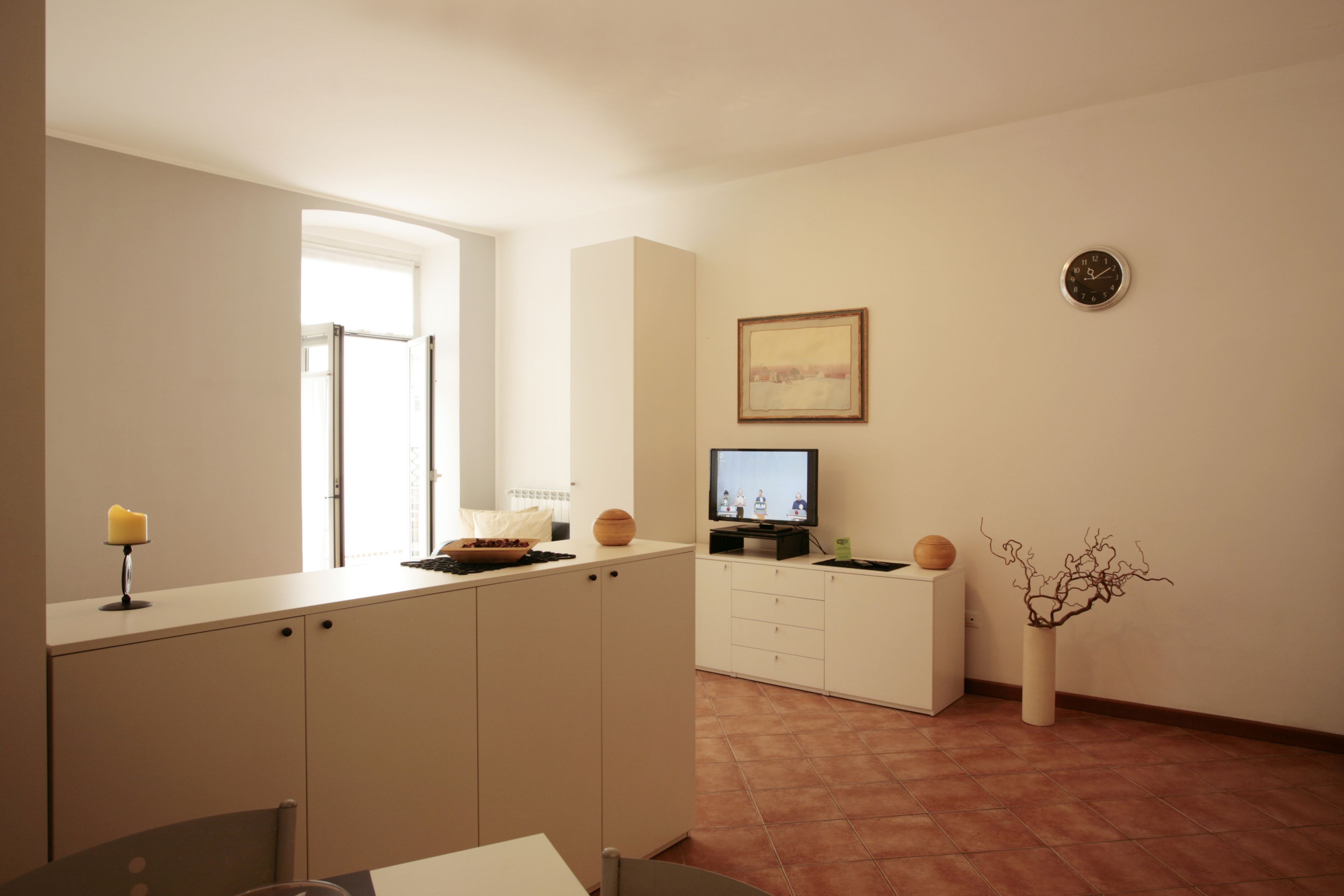 A modern kitchen with white cabinets, a dining area with a table and chairs, a TV on a stand, and a vase with branches on the wall.