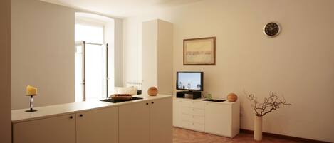 A modern kitchen with white cabinets, a dining area with a table and chairs, a TV on a stand, and a vase with branches on the wall.