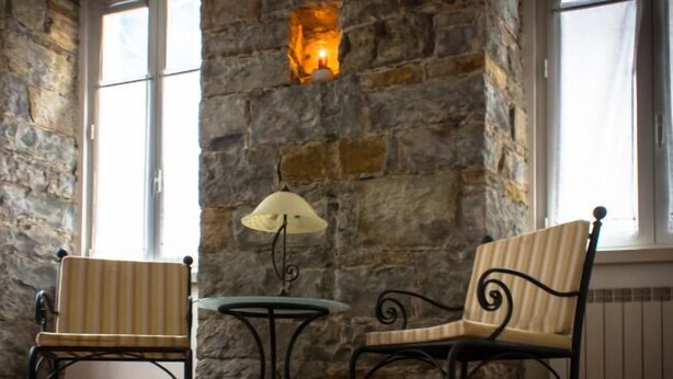 Habitación con paredes de piedra, dos sillones tapizados y una mesita, una lámpara de pared y un ventanal con cortinas.