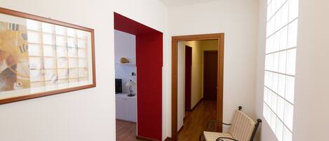 A hallway with a wooden floor, a red door, a framed picture on the wall, and a window on the right side.