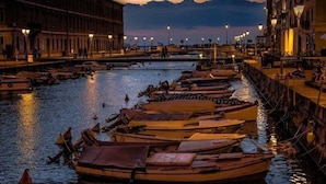 Una zona ribereña con barcos amarrados a lo largo de la orilla del río, edificios bordeando el canal y un cielo despejado al fondo.