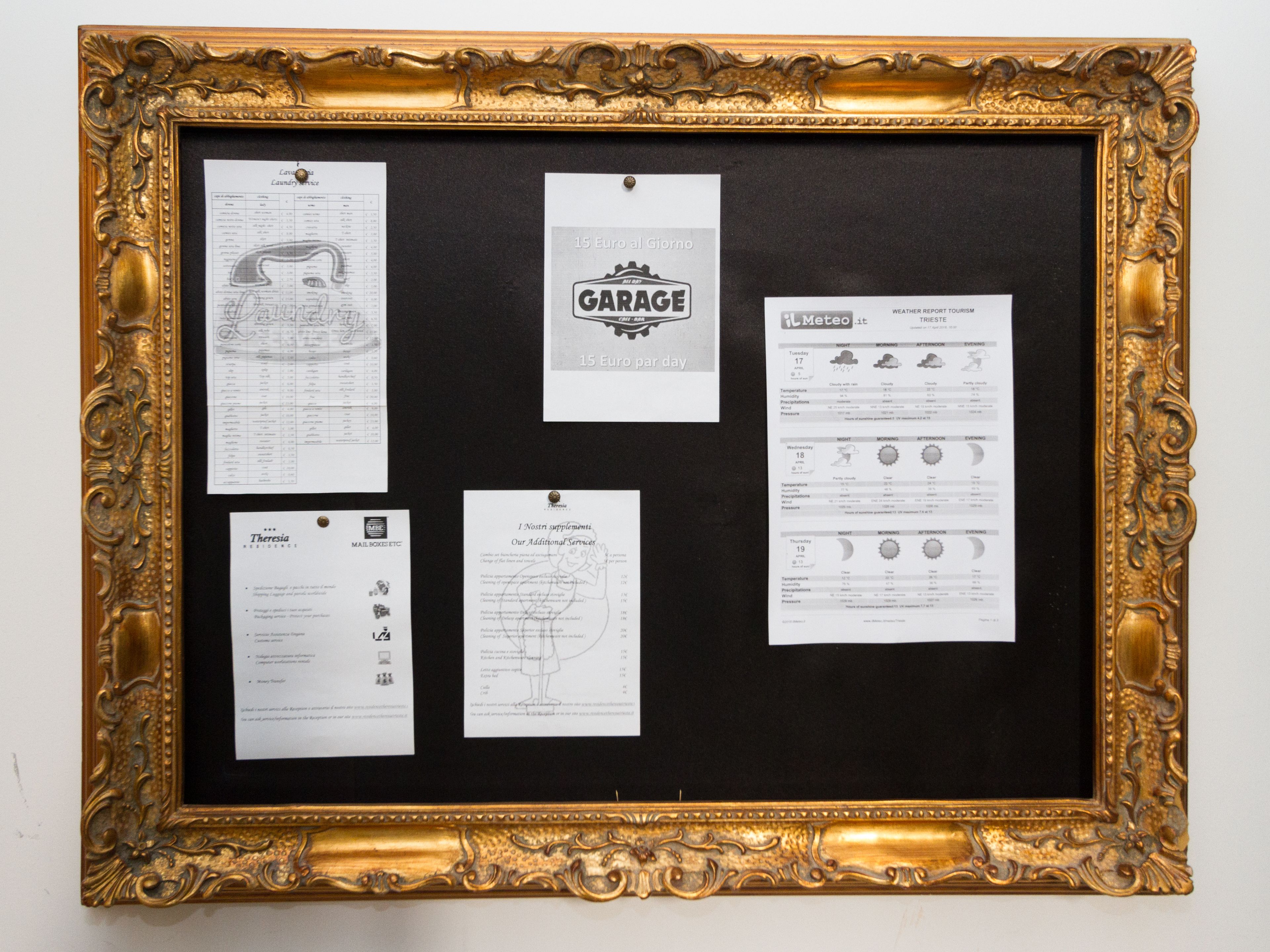 Un tablón de anuncios con varios documentos grapados, incluyendo un informe meteorológico y un cartel de garaje, en un marco dorado ornamentado.