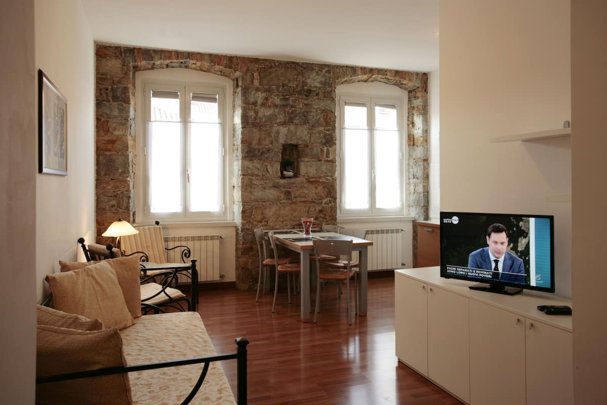 A cozy living room with a stone wall, a dining area with a table and chairs, and a television displaying a news program.