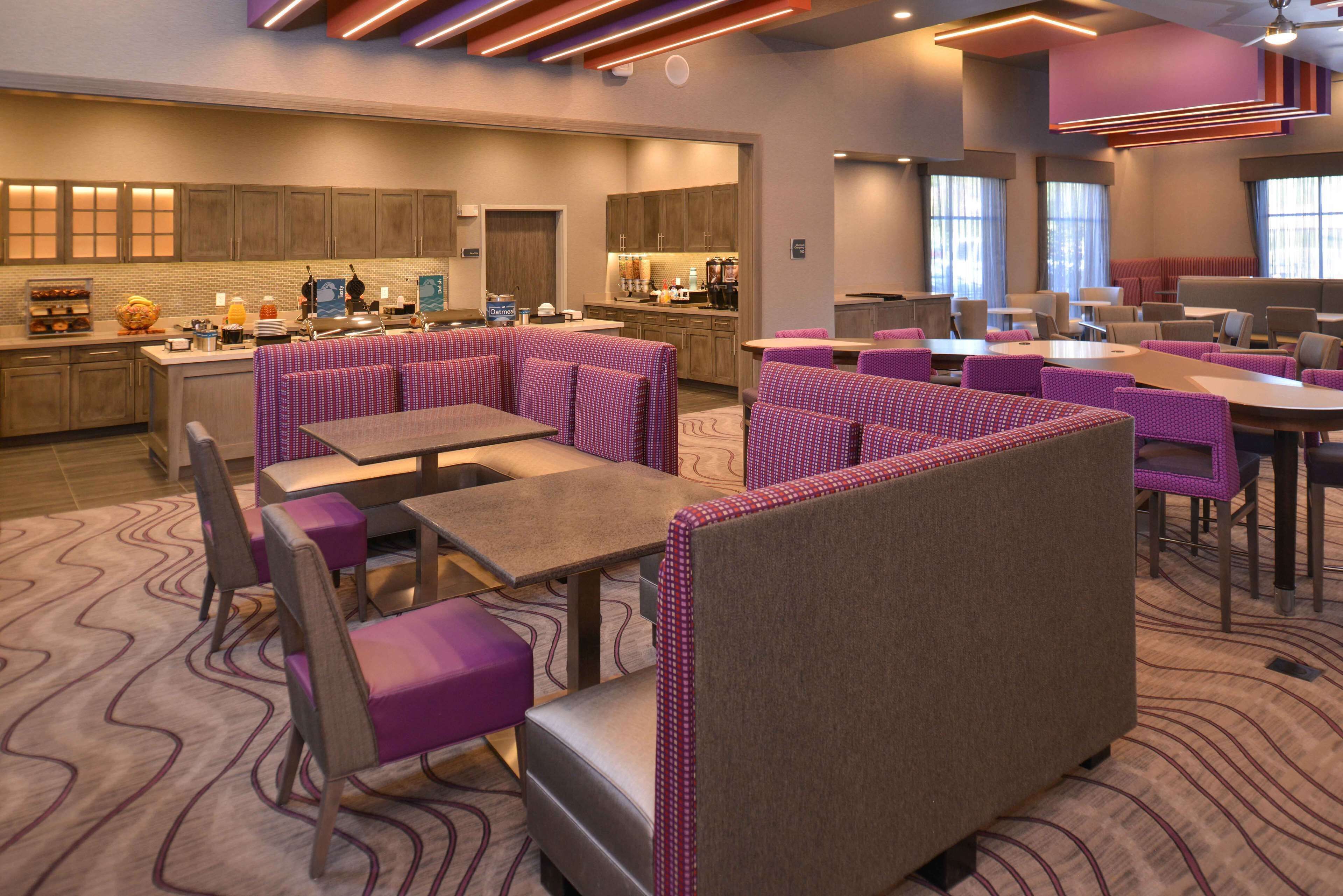 A modern hotel lobby with a central bar area, seating arrangements, and a kitchen in the background.