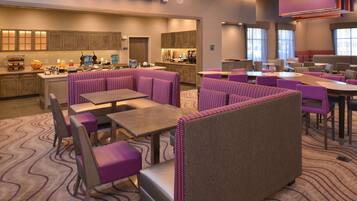 A modern hotel lobby with a central bar area, seating arrangements, and a kitchen in the background.