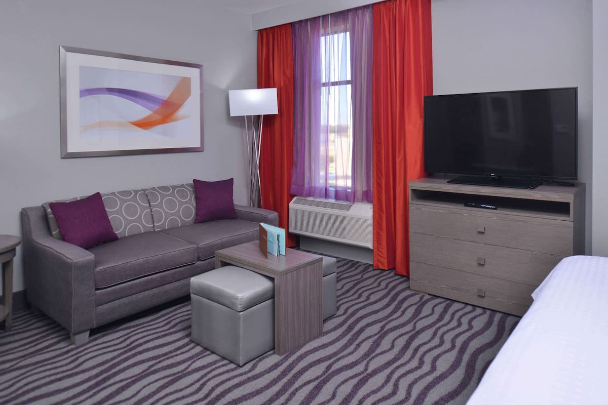 A hotel room with a grey sofa, a small ottoman, a flat-screen TV on a wooden stand, a bedside table, and a large window with red curtains.