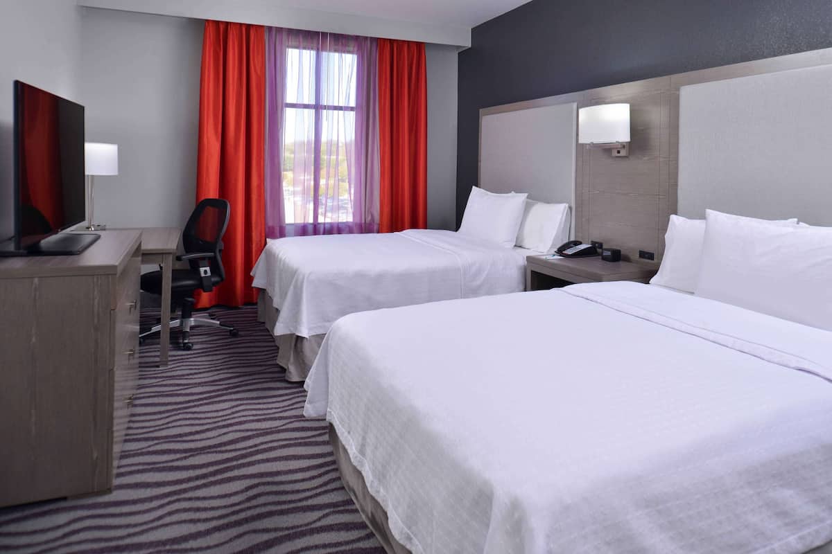 A hotel room with two beds, a desk, a chair, a TV, and a window with red curtains.