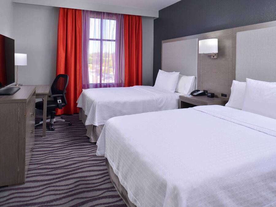 A hotel room with two beds, a desk, a chair, a TV, and a window with red curtains.