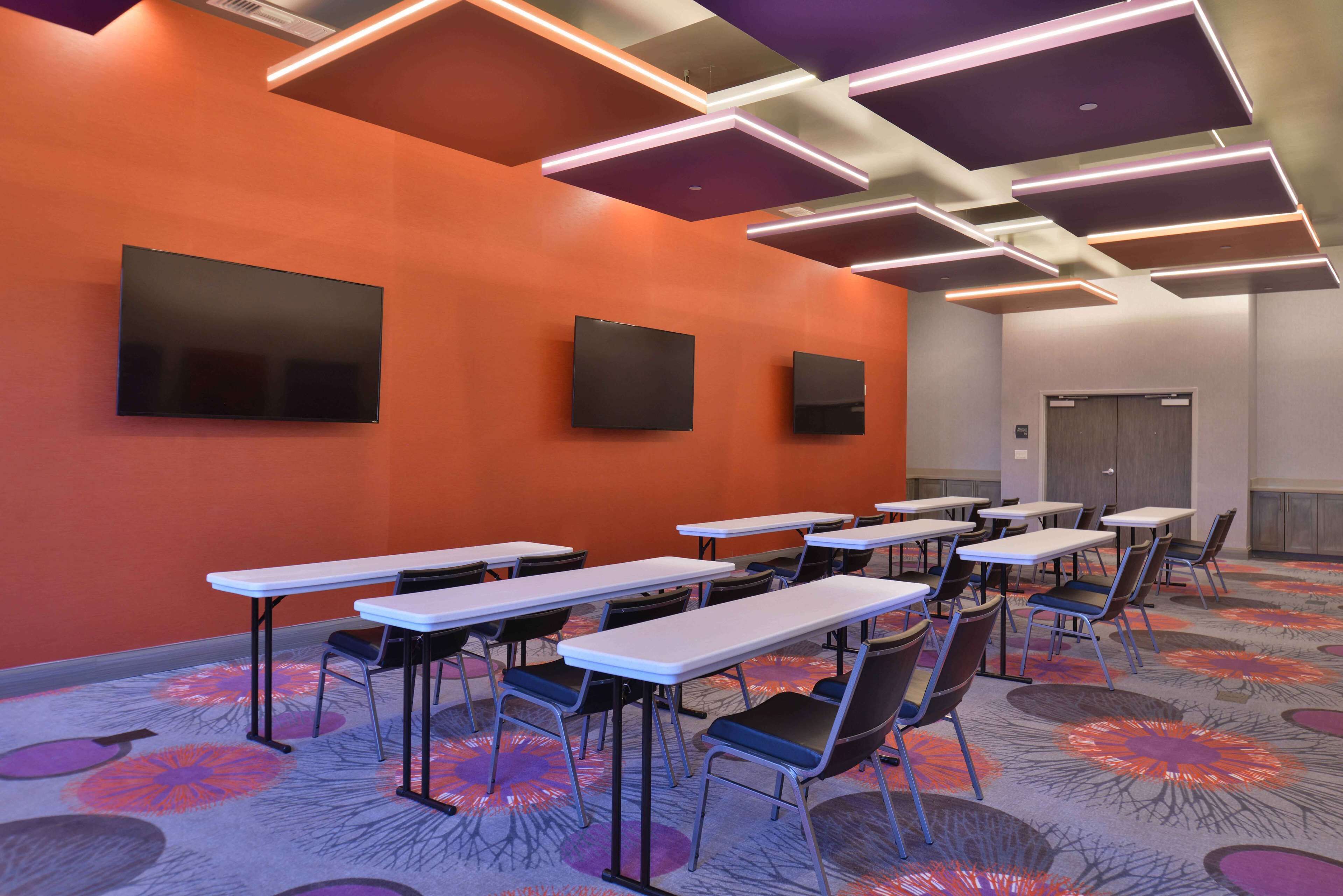 Une salle de conférence dotée de plusieurs tables et de chaises, d’un grand téléviseur à écran plat fixé au mur et d’un tapis à motifs.