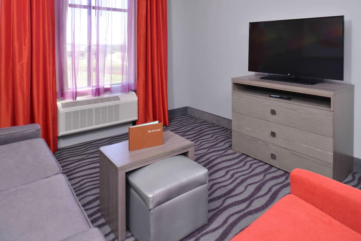 A hotel room with a flat-screen TV on a wooden stand, a grey sofa, a red armchair, a small table with a sign, and a zebra-patterned carpet.