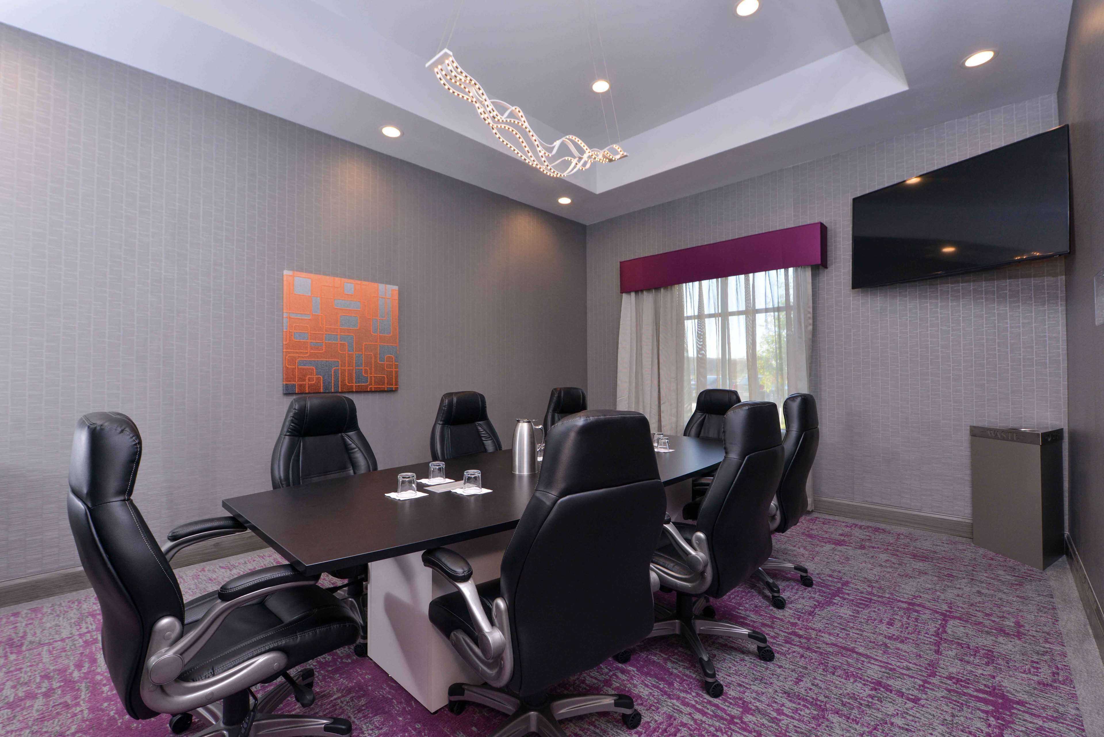 Un salón de conferencias con una larga mesa, sillas negras, un televisor de pantalla plana y una lámpara decorativa.