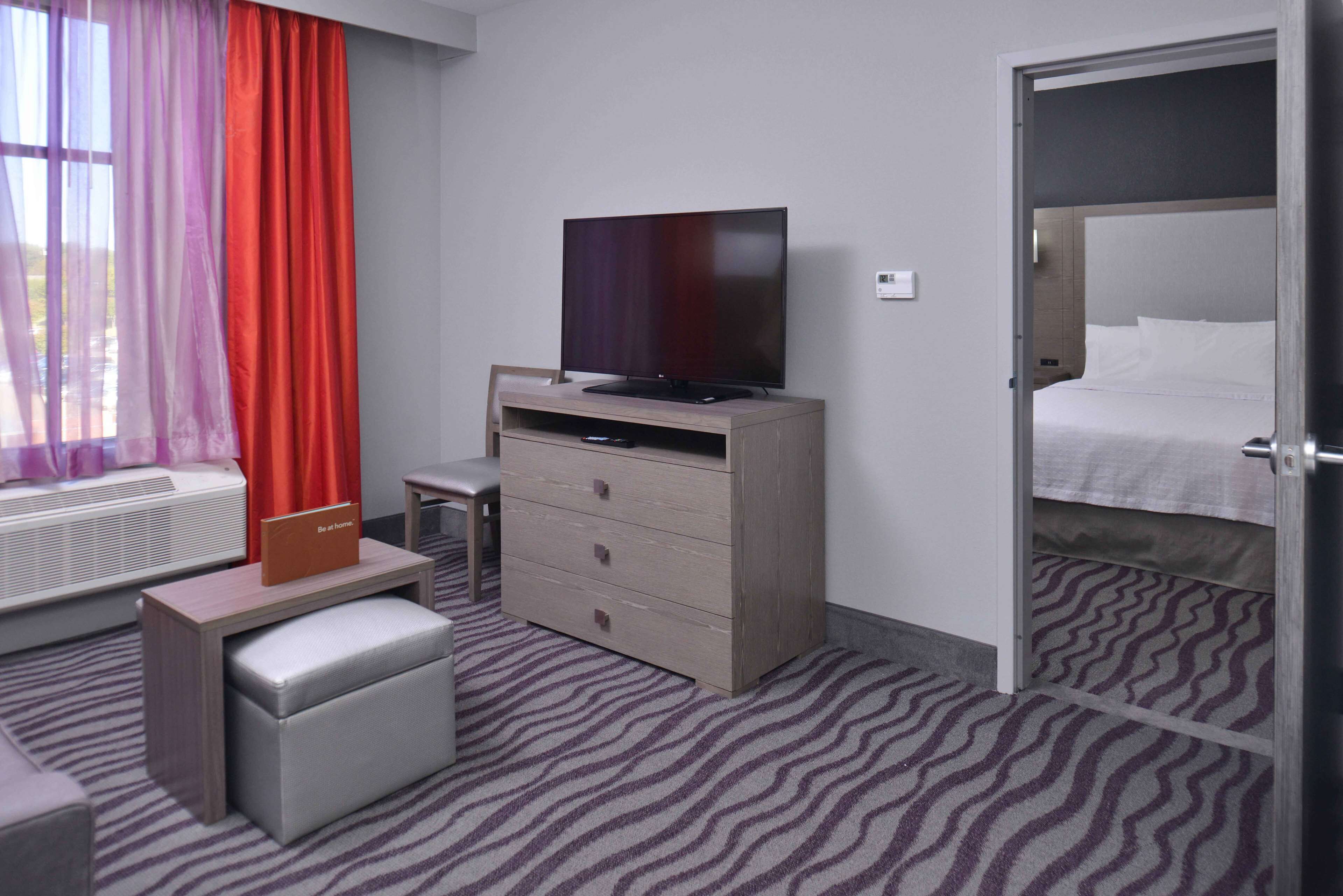 A hotel room with a flat-screen TV on a wooden dresser, a small table, a bench, a bed, and a window with red curtains.