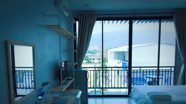 A hotel room with a bed, a desk, a chair, a TV, and a balcony with a view of buildings and mountains. - Sirin Loft Resident (Chiang Mai)
