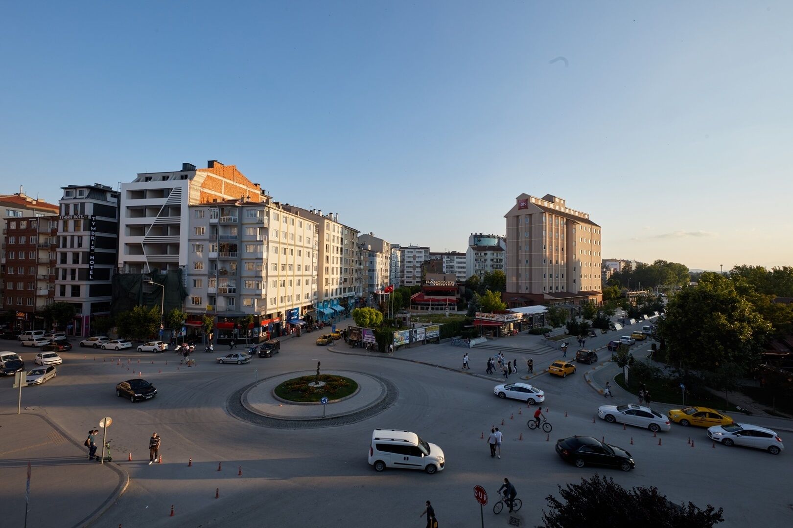 A city square with a roundabout, surrounded by multi-story buildings including a hotel and a bank.