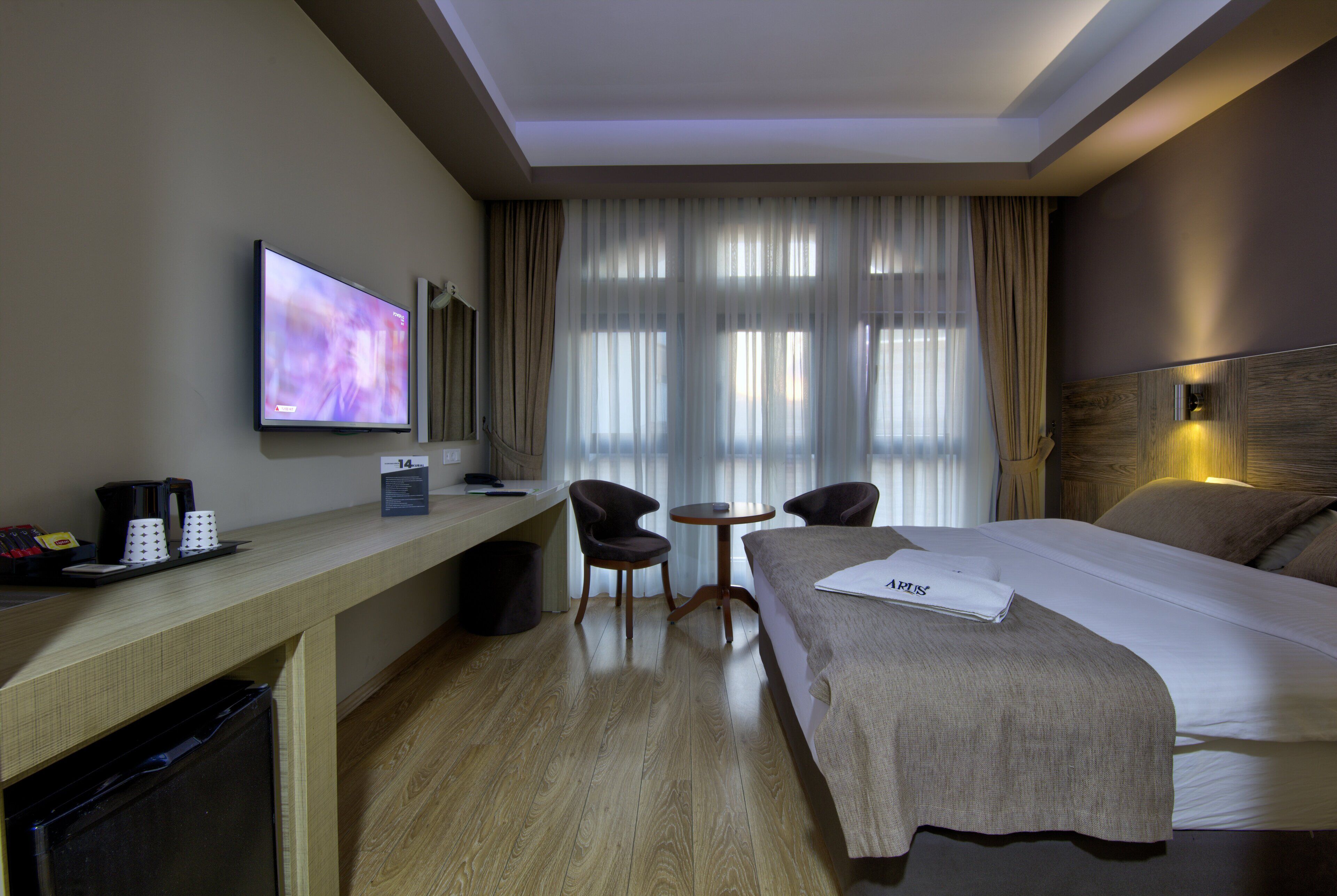 A hotel room with a bed, a desk with a TV, two chairs, a small table, and a trash bin.