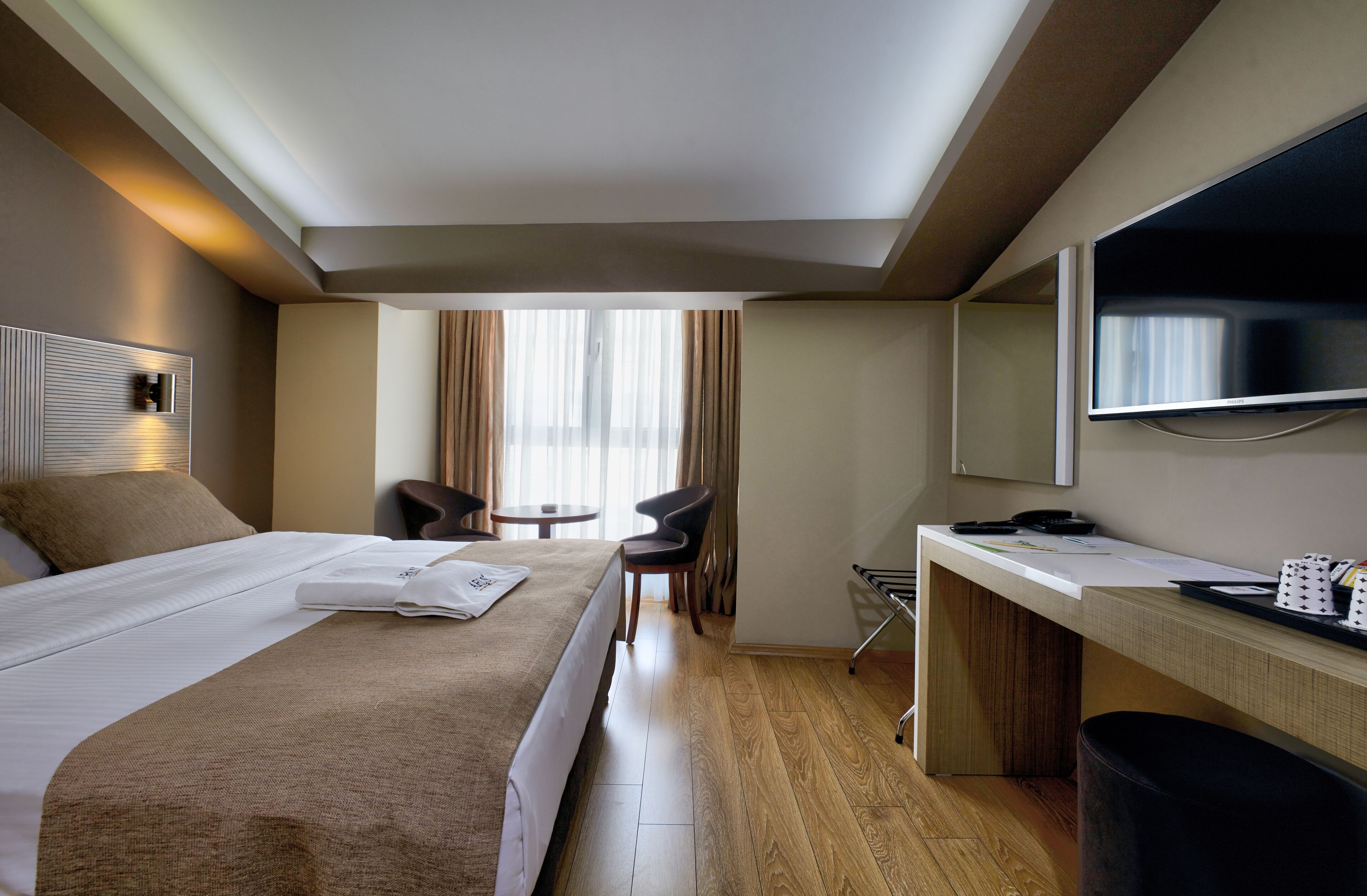 Quarto de hotel com cama, escrivaninha, cadeira, televisão e janela com cortinas.