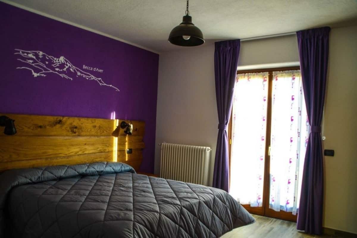 A bedroom with a large bed, purple walls, a wooden headboard, a radiator, and a window with purple curtains.