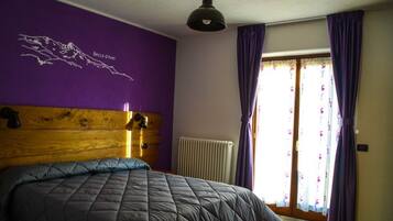 Un dormitorio con una cama grande, paredes moradas, cabecera de madera, un radiador y una ventana con cortinas moradas.